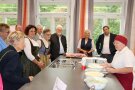 Große Küchenzeile, rechts Frau in Kochgewand mit Schüsseln, links und hinten schauen Gäste neugierig auf die Erklärende.