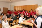 Frau steht rechts und begrüßt im Saal sitzende Personen.