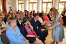 Frau in Dirndl mit Korb vor einem voll besetzten Sall mit Zuhörern verteilt in der ersten Reihe Sträußchen und Köstlichkeiten