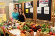 Zwei Frauen vor Tischen mit Lebensmitteln sowie Obst und Gemüse, im Hintergrund Schautafel.