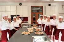 Neun Frauen mit Schürzen und Häubchen in Schulküche, auf dem Küchenblock viele Teller mit verschiedenen Schmalzgebäck-Varianten.