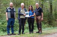 Vier Personen mit einer Broschüre stehen auf Waldweg, im Hintergrund Bäume und Sträucher.